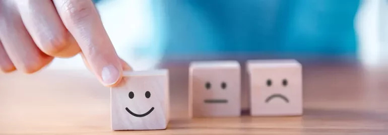 Fingers touching a wooden block with a smiley face, next to neutral and sad faces.