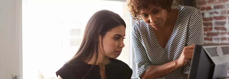 Two people working together at a computer in a well-lit room with brick walls.