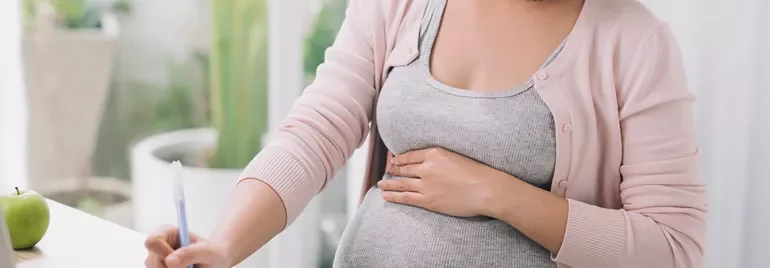 A person holding their pregnant belly while writing at a desk.