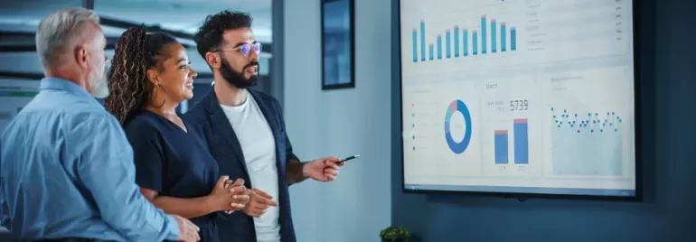Three people discussing statistics on a large screen in an office.