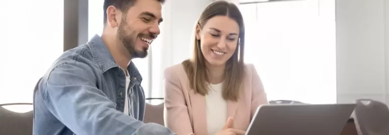 Two people are sitting at a table facing a laptop while smiling. 
