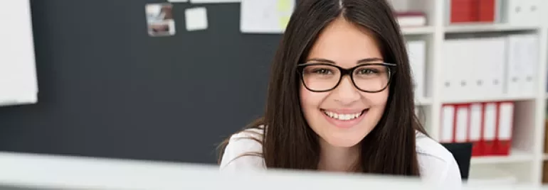 Person smiling in front of a computer with shelves and a board behind.