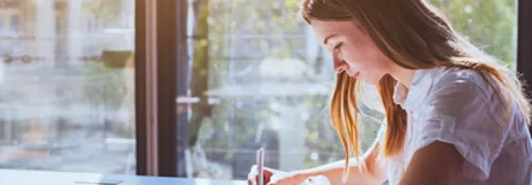 Person writing in a notebook at a sunlit desk.
