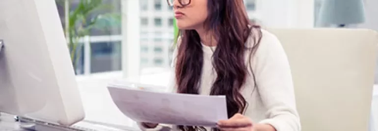 Person wearing glasses, focused on a document while sitting at a desk in front of a computer.