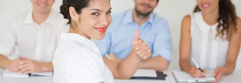 Person giving a thumbs up at a meeting with three people smiling in the background.
