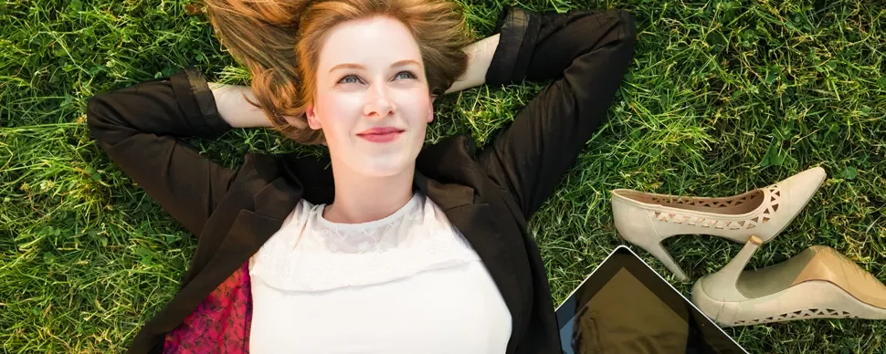Person lying on grass smiling, shoes and tablet nearby.