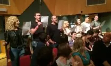 People singing in a recording studio, holding sheets of paper.