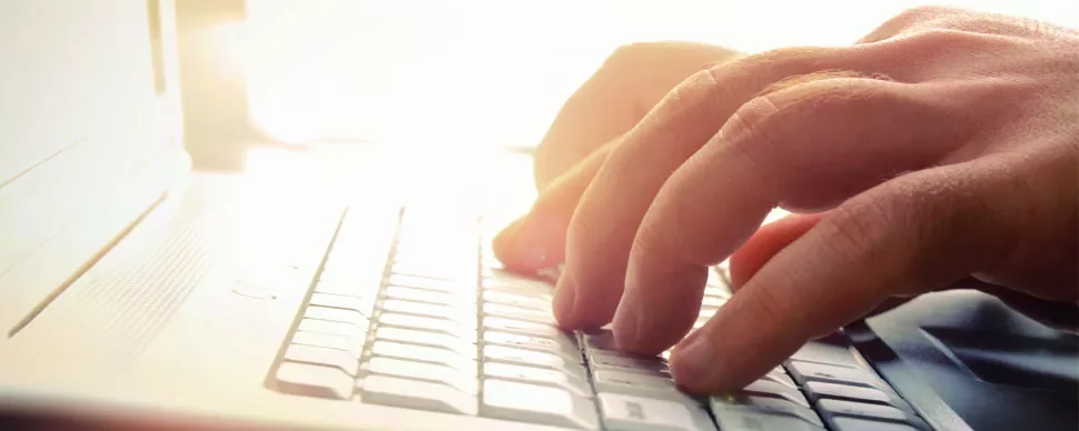 Hands typing on a laptop keyboard with sunlight in the background.