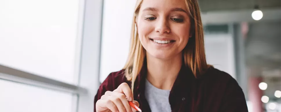 Person smiling while writing with a pen at a table near a window.
