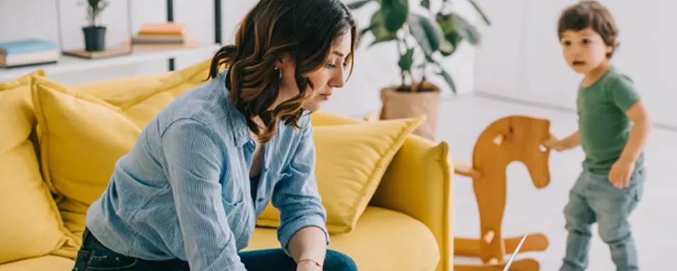 Person sitting on yellow sofa, child playing with wooden horse in living room.