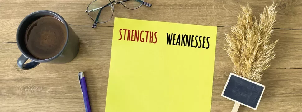 Yellow paper labeled strengths and weaknesses on a wooden desk with coffee, glasses, pen, and wheat.