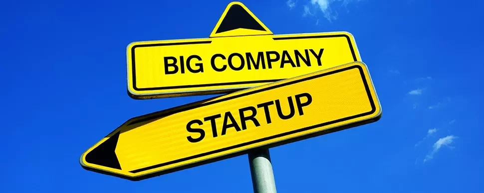 Two yellow directional signs pointing to big company and startup against blue sky.