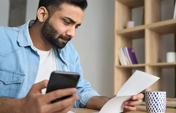 Person reading a paper while holding a phone, with a mug on the table.