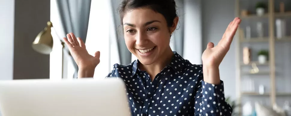 Person smiling and looking at a laptop, raising hands in excitement.