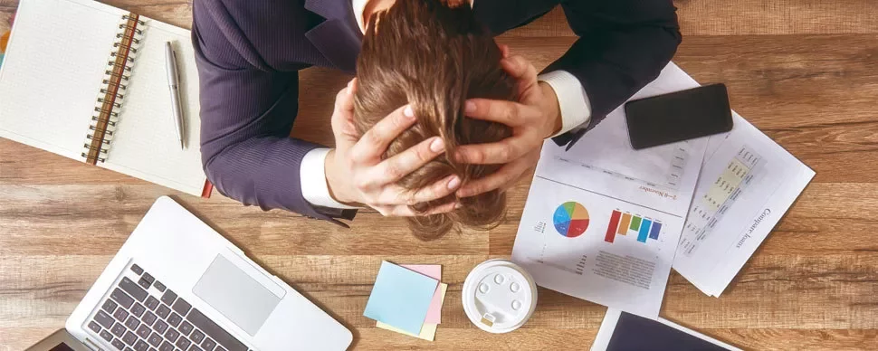 Person at desk with head in hands, surrounded by documents, a laptop, and coffee.