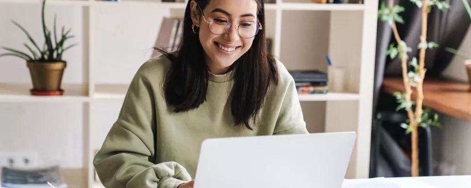 Smiling person using a laptop at a desk with shelves in the background.