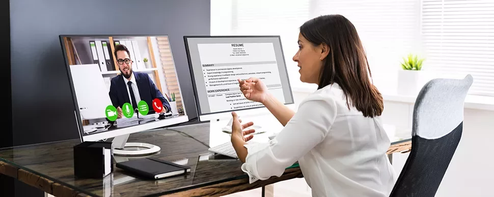 Person in a video call interview at a desk with dual monitors displaying resume and video screen.