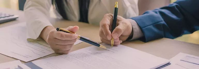 Two people reviewing documents with pens on a table.