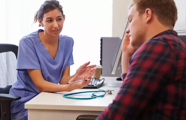 A medical professional talks to a person at a desk with a stethoscope nearby.