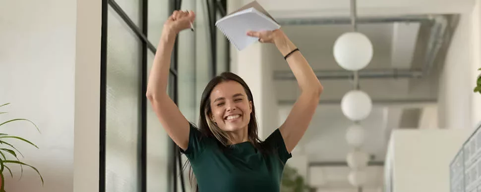 Person celebrating in an office, holding a notebook with arms raised.