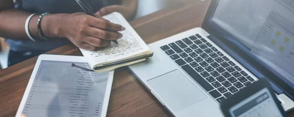 Person writing in notebook next to a laptop and tablet.