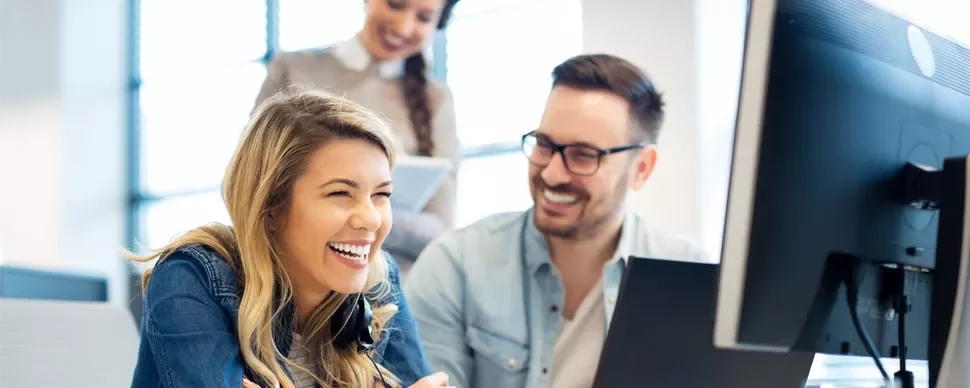 Three people smiling and working together at a computer desk.