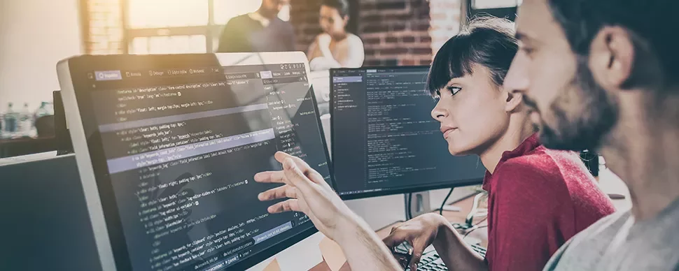 Two people collaborating on programming at dual monitors in an office.