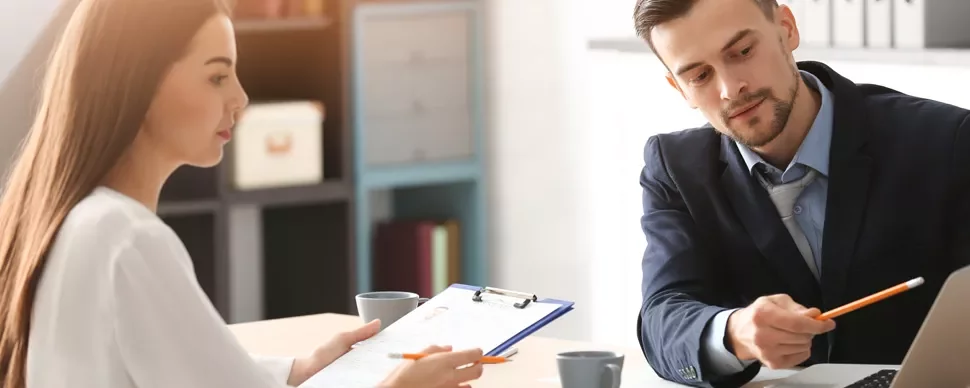 Two people discussing documents at a table, one holding a clipboard.