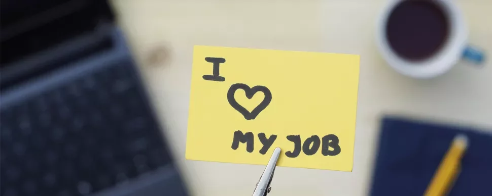 Yellow note with 'I love my job' held over a blurred laptop and coffee cup.