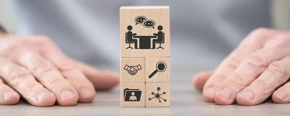 Wooden blocks with icons of communication, handshake, magnifying glass, document, and network. Hands visible.