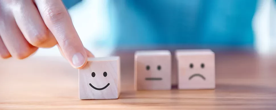 Fingers touching a wooden block with a smiley face, next to neutral and sad faces.
