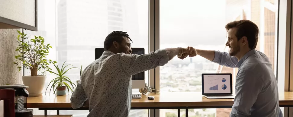 Two colleagues smiling and fist-bumping in an office with charts on a laptop.