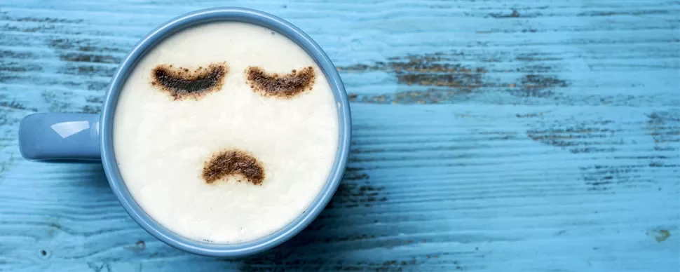 Coffee cup with a sad face design in the milk foam on blue table.