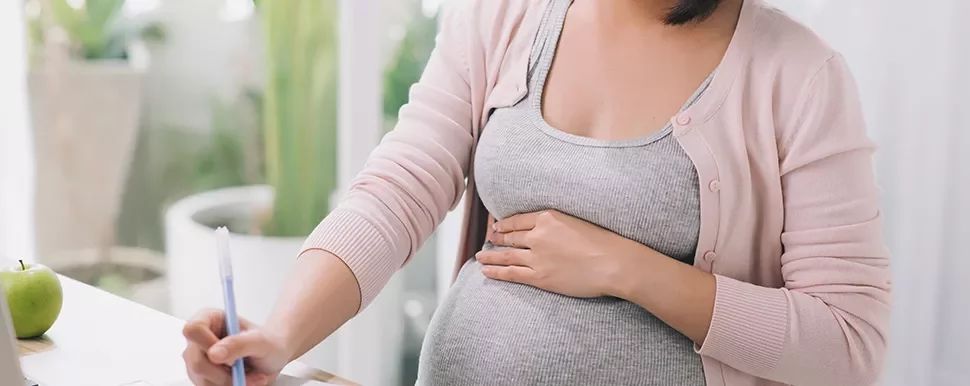 A person holding their pregnant belly while writing at a desk.