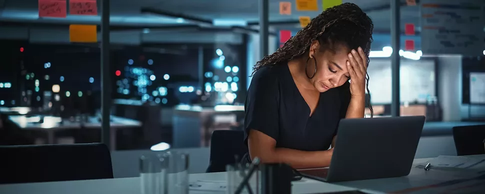 Person sitting at a desk with a laptop, appearing thoughtful in a dimly lit office.