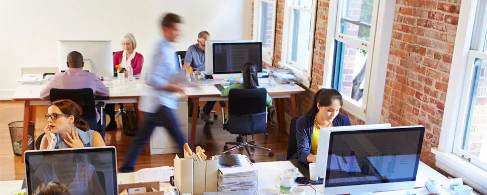 Open office with people working at desks, some using computers, and one person walking.