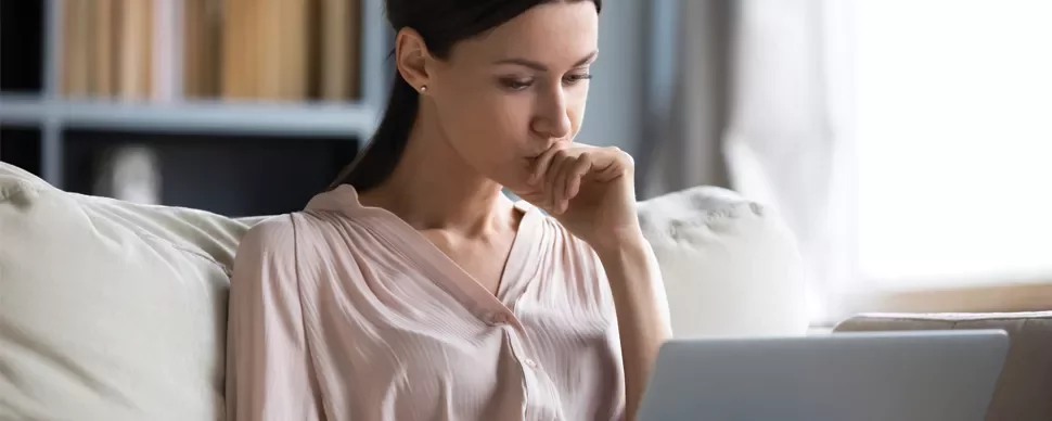 Person seated on a couch, looking at a laptop screen with a thoughtful expression.
