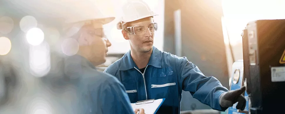 Two workers in protective gear discuss machine operations in a factory setting.