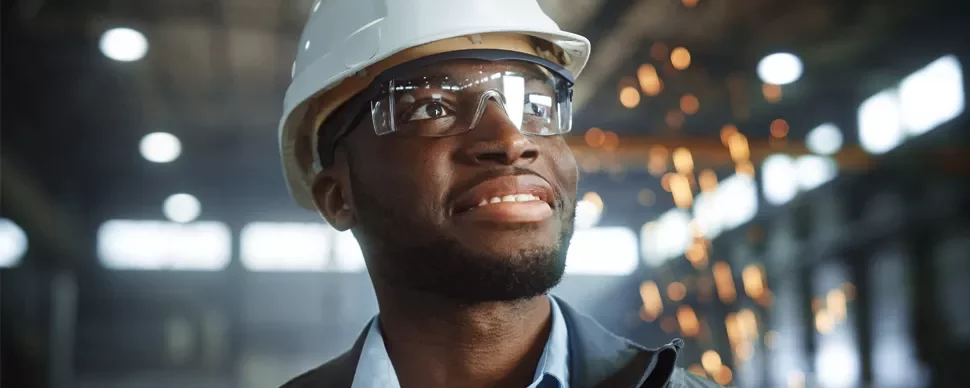 Person wearing a hard hat and safety glasses smiles in a workshop with sparks in the background.