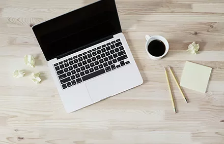 A laptop, coffee cup, pencils, and crumpled papers on a wooden desk.
