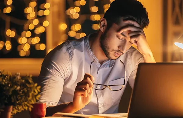 Person sitting at a desk, holding glasses, looking at a laptop.