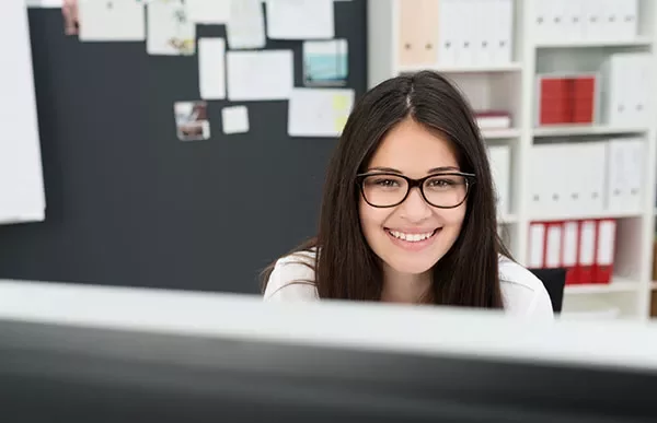 Person smiling in front of a computer with shelves and a board behind.