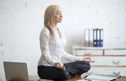 Person meditating on a desk in a serene office setting with a laptop and notebooks nearby.