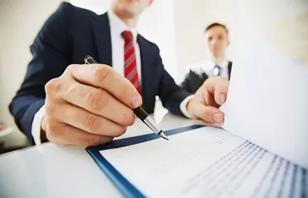 Person signing a document at a desk with another person in the background.