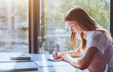 Person writing in a notebook at a sunlit desk.