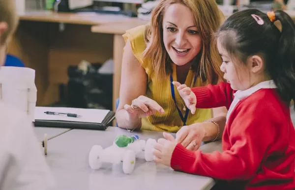 Teacher smiles while helping a student with a project at a desk.