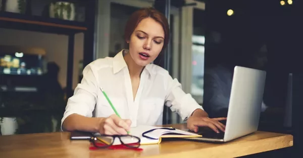 Person writing notes at a desk with an open laptop.