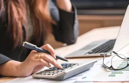 Person using calculator at desk with laptop and glasses.