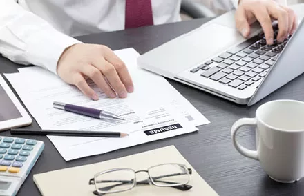 Person working on a laptop with paperwork and a coffee mug on the desk.