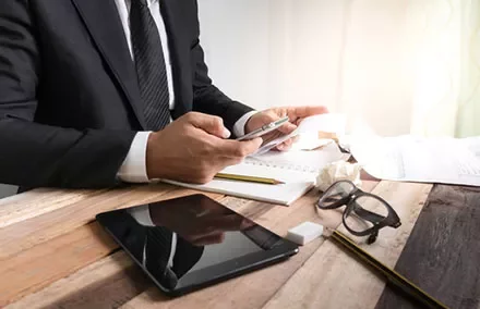 Person in suit using smartphone at a desk with a tablet, glasses, and papers.